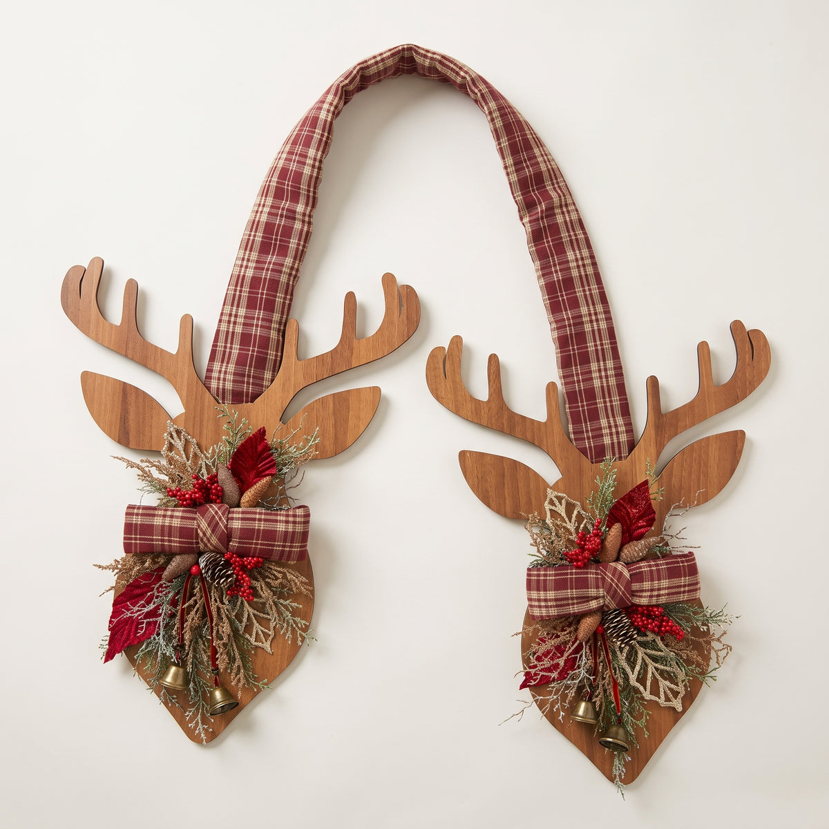 Handcrafted two-sided wooden deer holiday door hanger featuring a festive red plaid tartan ribbon, frosted pine branches, pinecones, berries, and a golden bell, against a white background