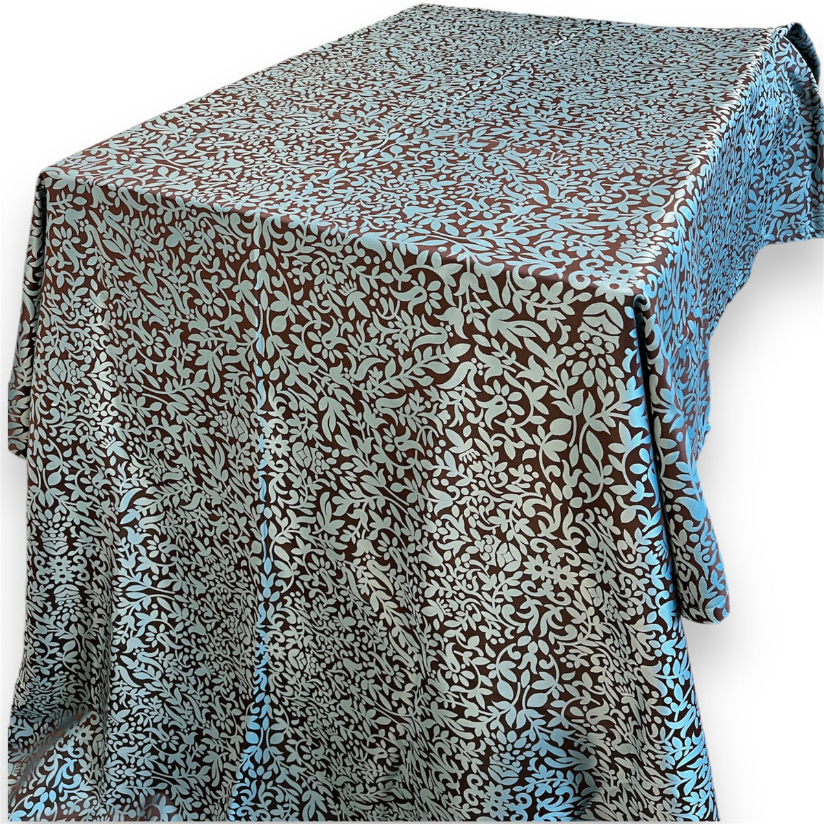 Rectangular tablecloth with a dense light blue floral and leaf vine pattern on a dark brown background draped over a table against a white background.