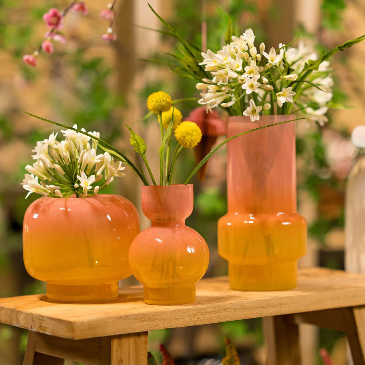 Three pink and orange gradient glass vases of various shapes holding fresh white and yellow floral stems on a rustic wooden bench.