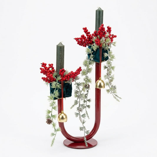 A decorative U-shaped candle holder with a glossy dark red base and curved arms, holding two dark green taper candles. The arrangement is decorated with red berry sprigs, white pine branches, small pinecones, and hanging gold bells, set against a plain white background.