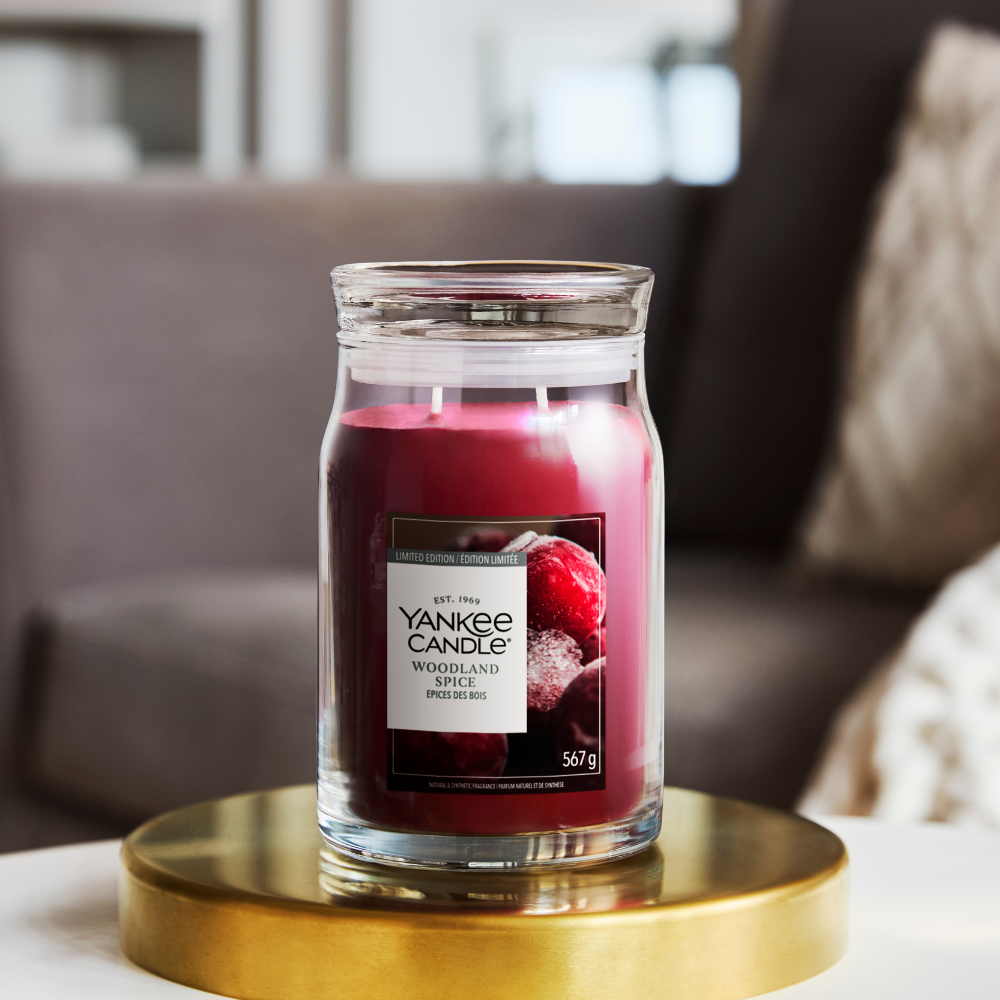 A Yankee Candle 'Woodland Spice' scented candle in a clear glass jar, resting on a round gold tray. The background shows a softly blurred living room setting with a gray sofa and a textured throw pillow.
