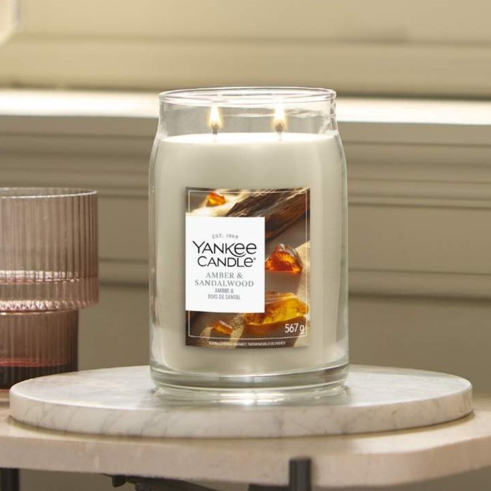 A Yankee Candle scented soy wax candle in a clear glass jar with two lit wicks, placed on a round marble coaster. The label reads 'Yankee Candle Amber & Sandalwood'.