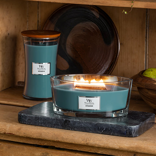 Lit WoodWick Antiquarium Ellipse Jar Candle glowing on a dark marble slab, alongside a tall WoodWick Hourglass Jar Candle on a rustic wooden shelf.