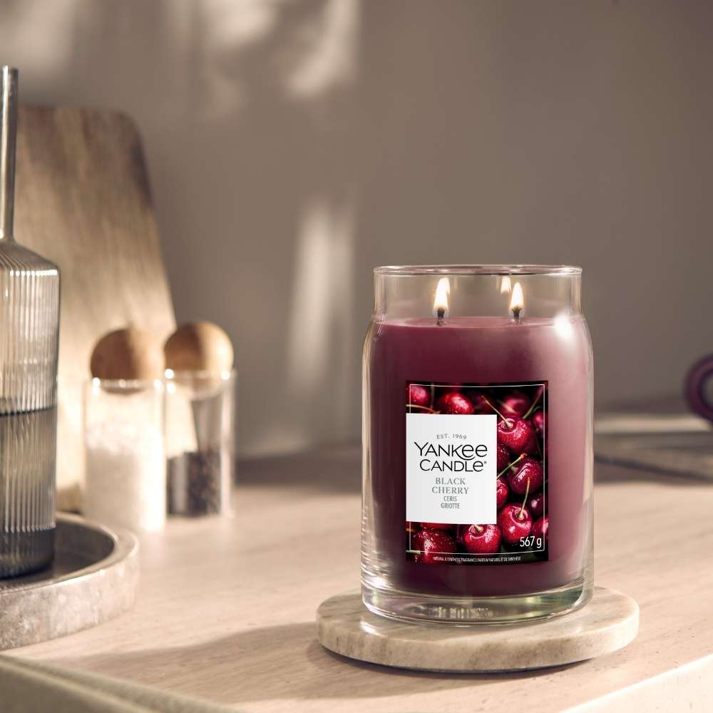 A Yankee Candle All Over Cherry scented candle in a clear glass jar with two wicks, sitting on a round marble coaster on a wooden table in a softly lit room.