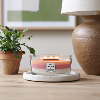 A lit three-layered scented candle in a clear glass jar sits on a white marble coaster on a wooden table. The candle has peach, pink, and white layers, with a white label that reads 'Ww TRILOGY'.
