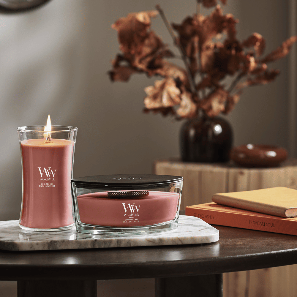 WoodWick Cinnamon Chai scented candles displayed on a wooden table, including an hourglass glass jar candle with a lit wooden flame and an ellipse vessel candle resting on a marble trivet alongside books and dried floral arrangements.