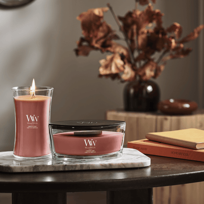 WoodWick Cinnamon Chai scented candles displayed on a wooden table, including an hourglass glass jar candle with a lit wooden flame and an ellipse vessel candle resting on a marble trivet alongside books and dried floral arrangements.