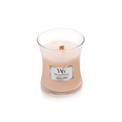 A WoodWick Coastal Sunset scented candle in a clear glass jar with a light peach-pink wax and a single wooden wick, set against a plain white background.