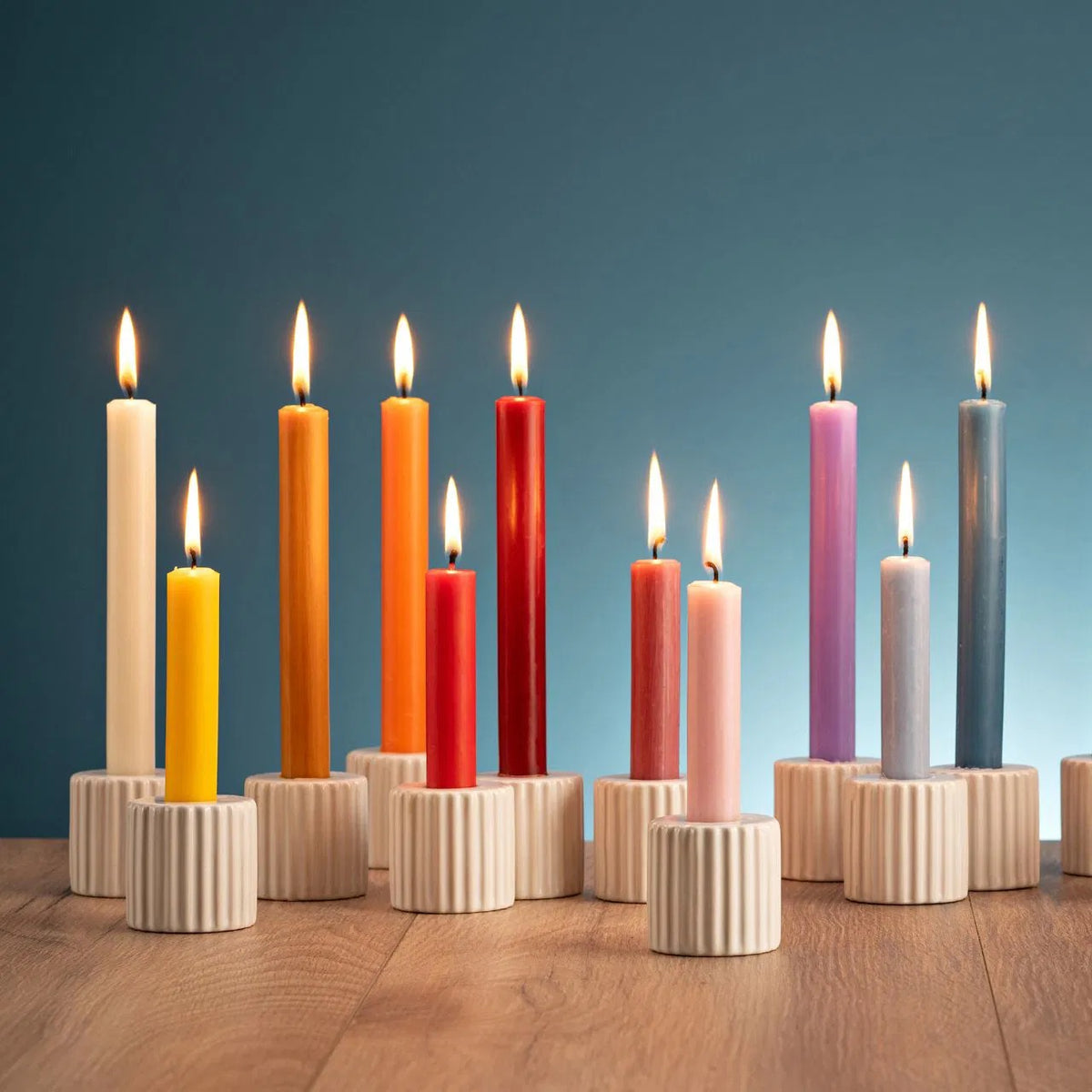 Row of colorful lit taper candles in beige ribbed ceramic holders arranged on a wooden surface against a teal background.