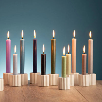 Row of colorful lit taper candles in beige ribbed ceramic holders arranged on a wooden surface against a teal background.
