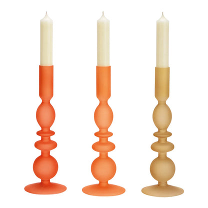 Three glass candle holders in red-orange, orange, and gold-beige colors, each with a candle lit on top.