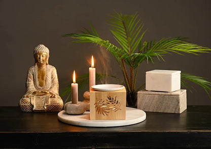 Serene golden Buddha figurine styled on a dark table alongside burning taupe candles, an essential oil burner, marble containers, and green palm leaves.