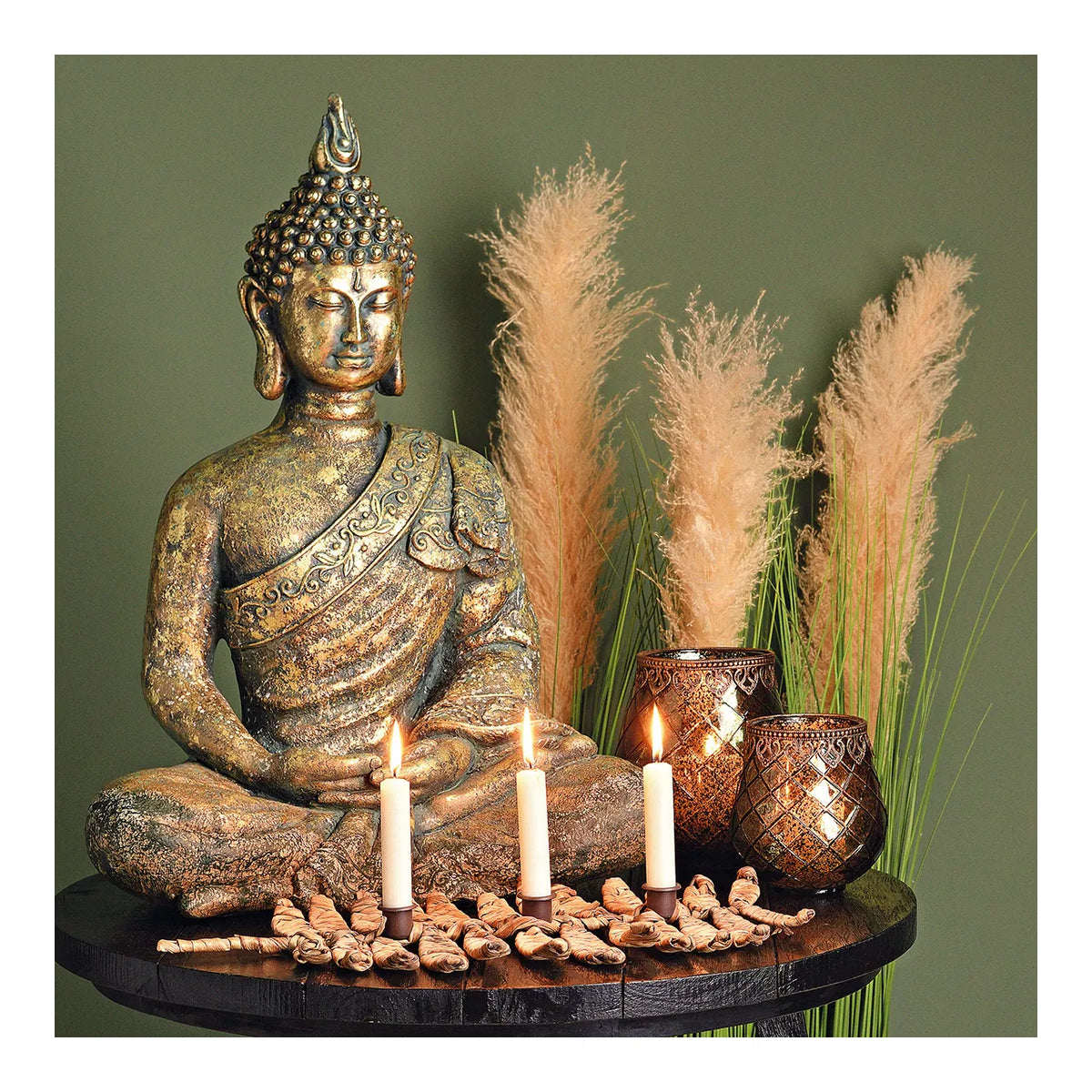 Golden serene Buddha statue seated with lit ivory stick candles and patterned glass votives on a dark round table against pampas grass.