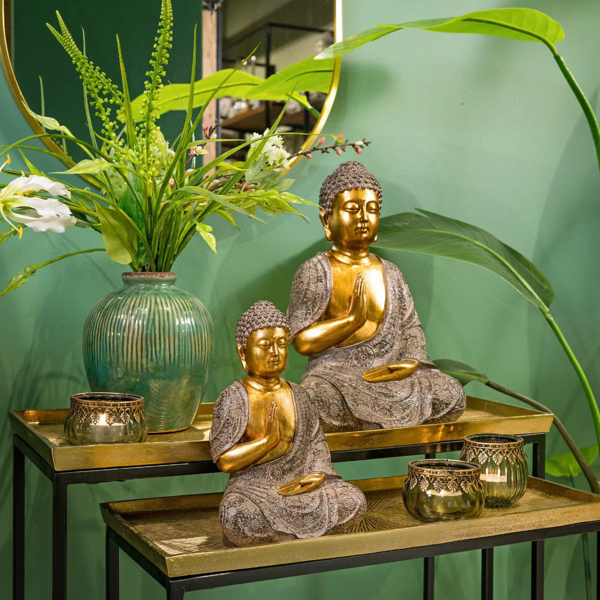 Green glass tealight candle holders displayed on tiered gold accent tables alongside Buddha statues and vibrant green plants.
