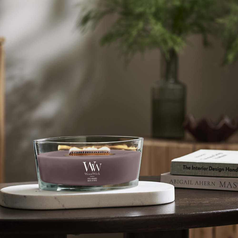 WoodWick Iris Woods ellipse candle in a clear glass jar with an open lid against decorative backround