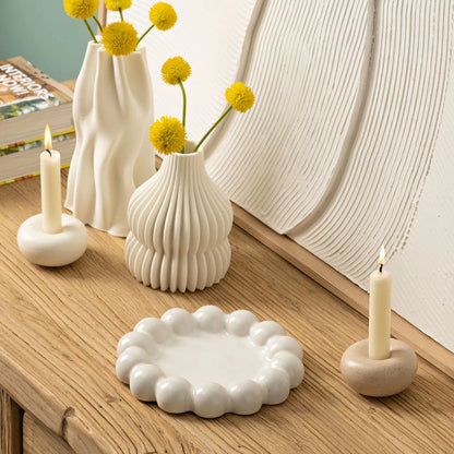 Two lit ivory stick candles in low ceramic bases styled on a wooden tabletop with ribbed white ceramic vases, yellow Billy Button flowers, and a scalloped white tray.