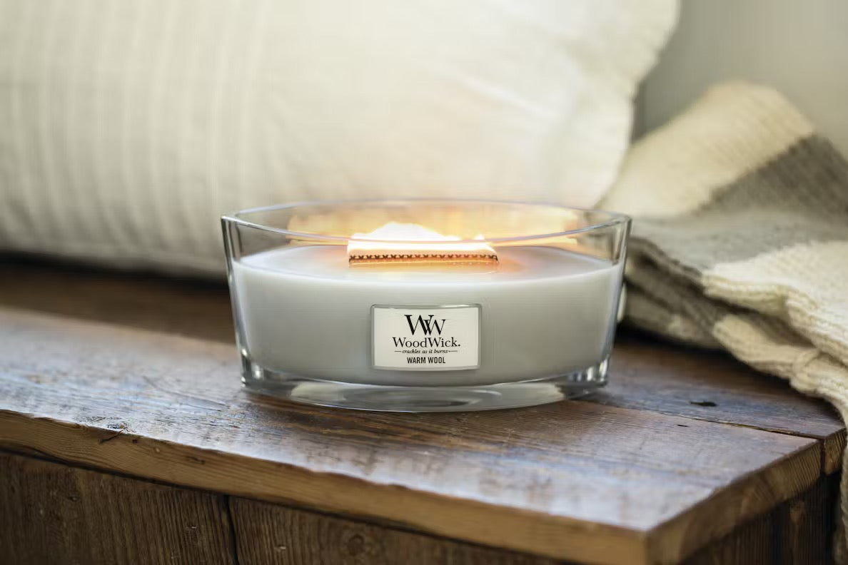 Lit WoodWick Warm Wool Ellipse Jar Candle glowing on a rustic wooden table, with soft white pillows and a folded throw blanket in the background.