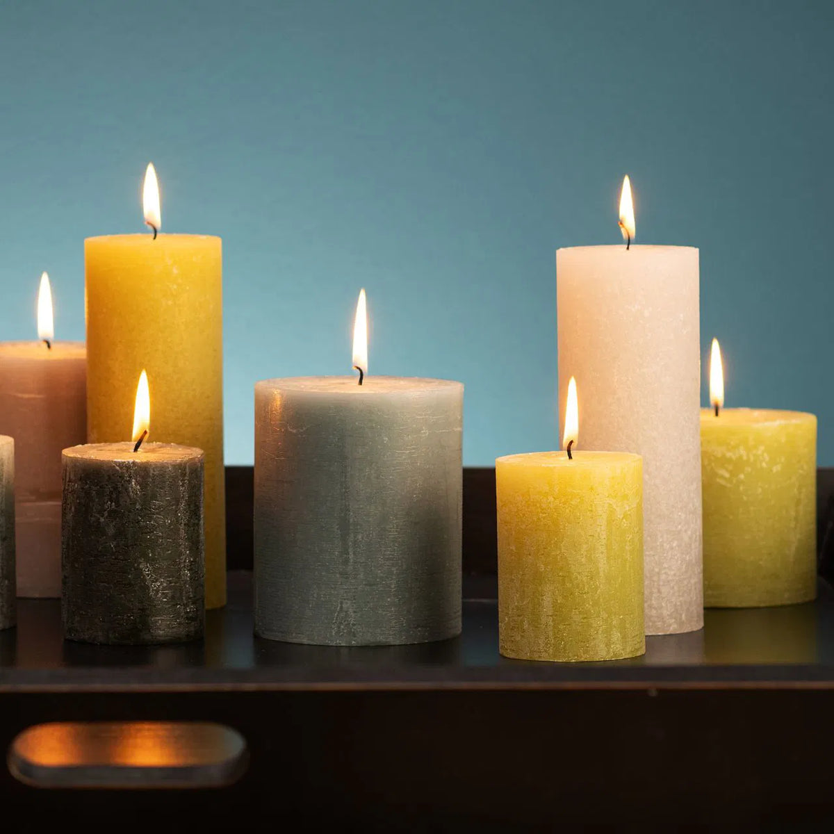 Collection of lit rustic textured pillar candles in various sizes, colors, and shades of green, yellow, beige, and grey.