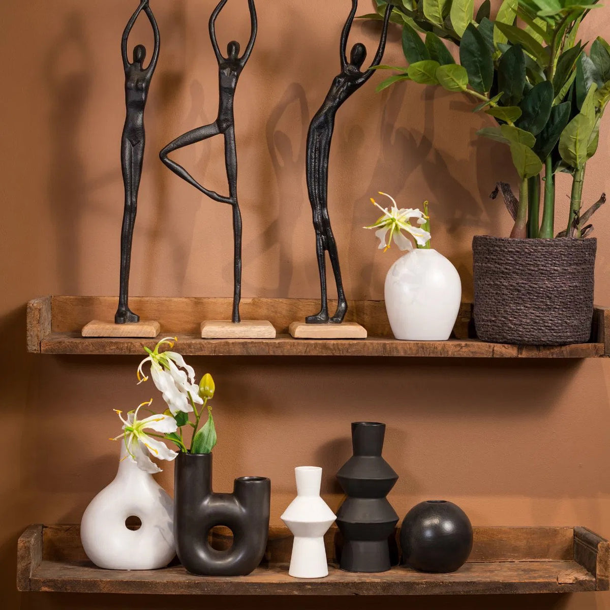 Three black metal human stand figures posed on wooden bases, styled on rustic wooden shelves with modern black and white ceramic vases and potted plants.