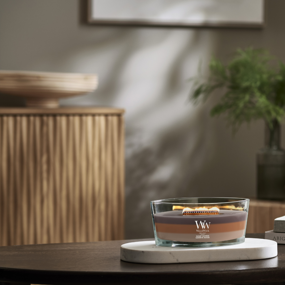 WoodWick Radiant Cashmere trilogy ellipse candle in a clear glass jar with an open lid against decorative backround.
