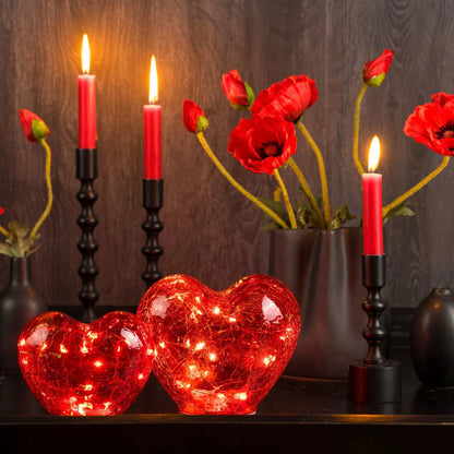 Two glowing red glass heart-shaped ornaments with warm fairy LED lights inside, arranged with red candles and poppies.