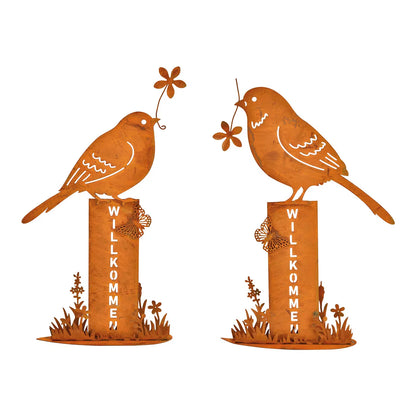 Two metal bird stands with a rusty finish, each featuring a bird holding a flower, mounted on posts with the word 'WELCOME' on them.