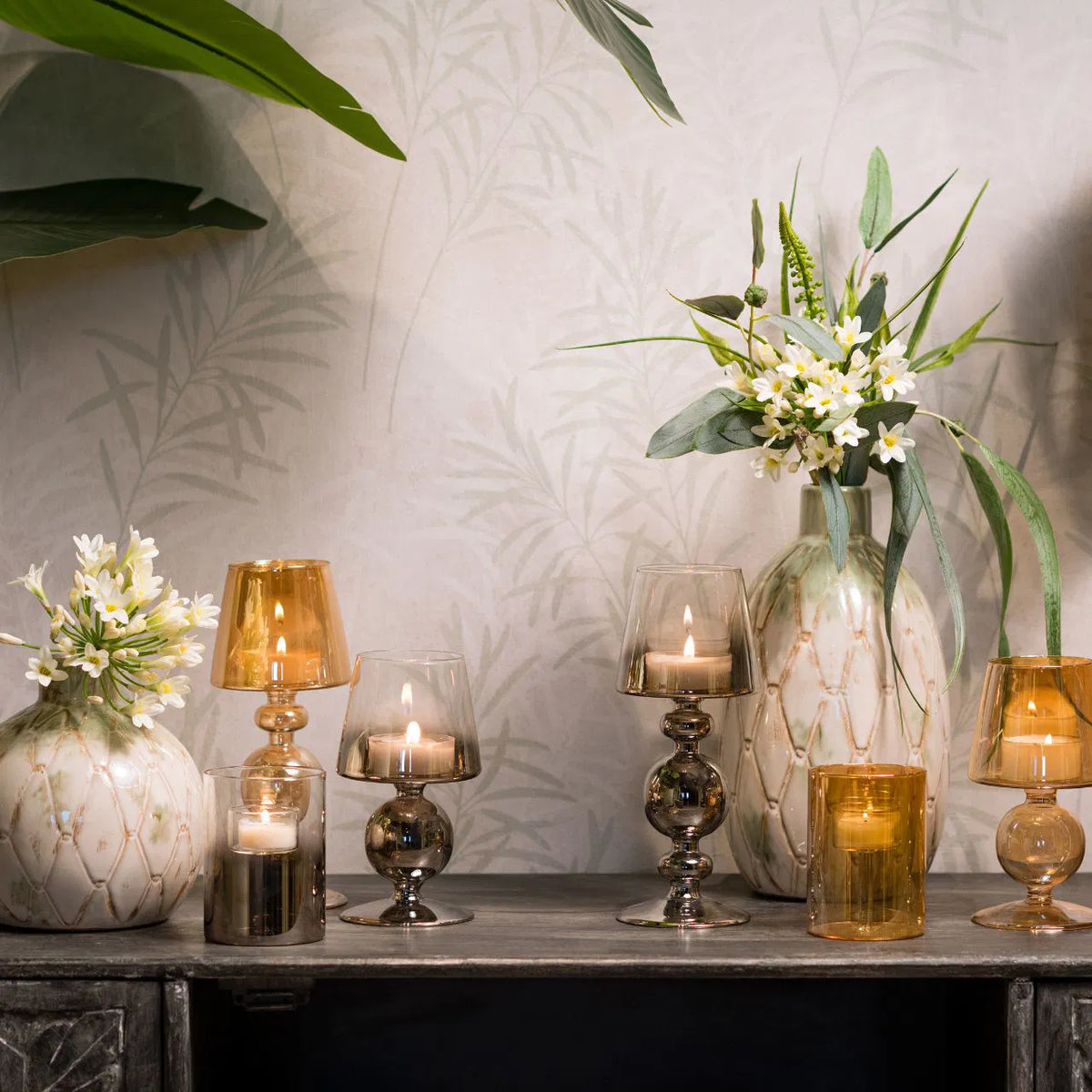 Assorted amber cognac and dark silver glass candle holders with lit candles, styled on a console table alongside textured ceramic vases and greenery.