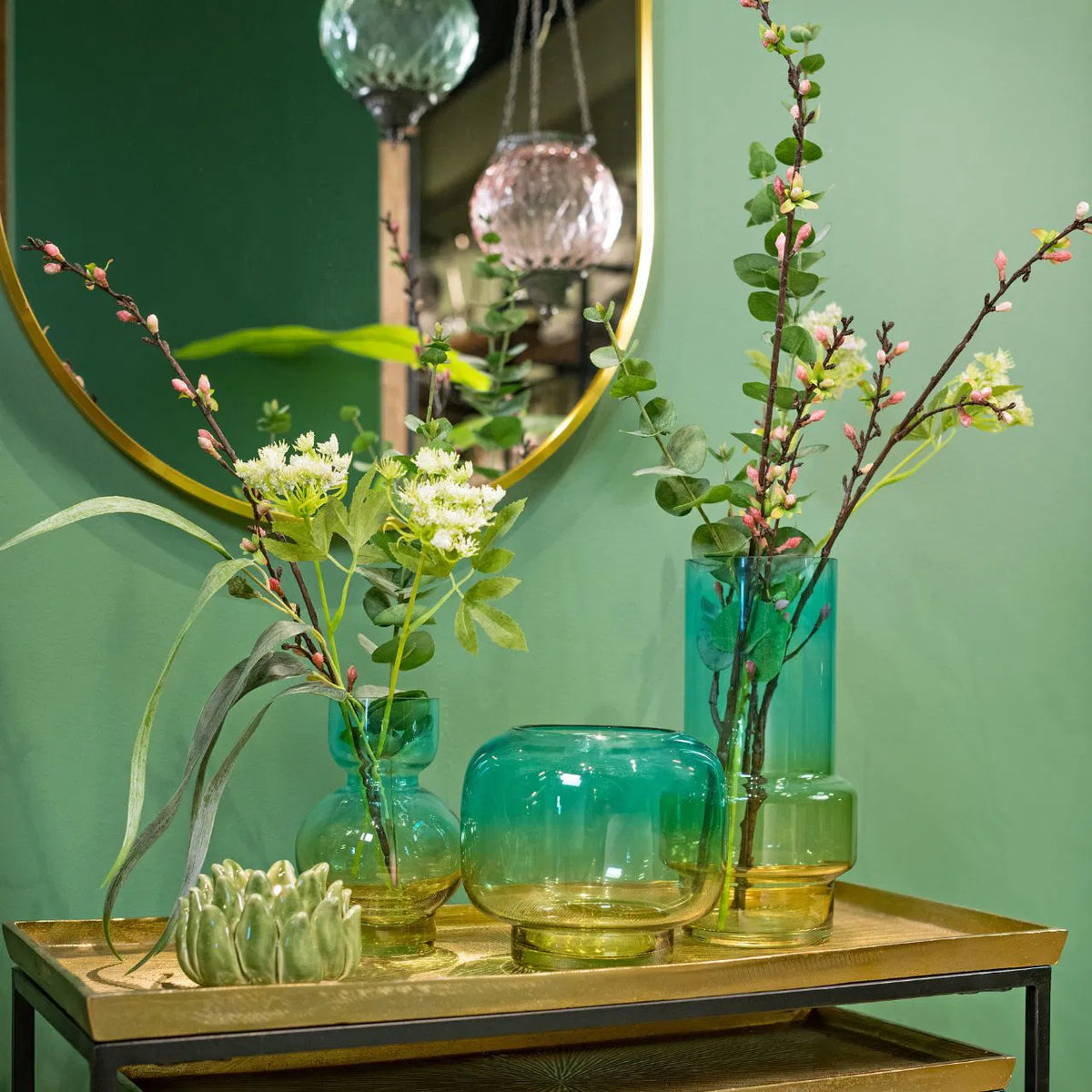 Collection of blue and green glass vases in various shapes filled with leafy floral stems on a metallic tray table