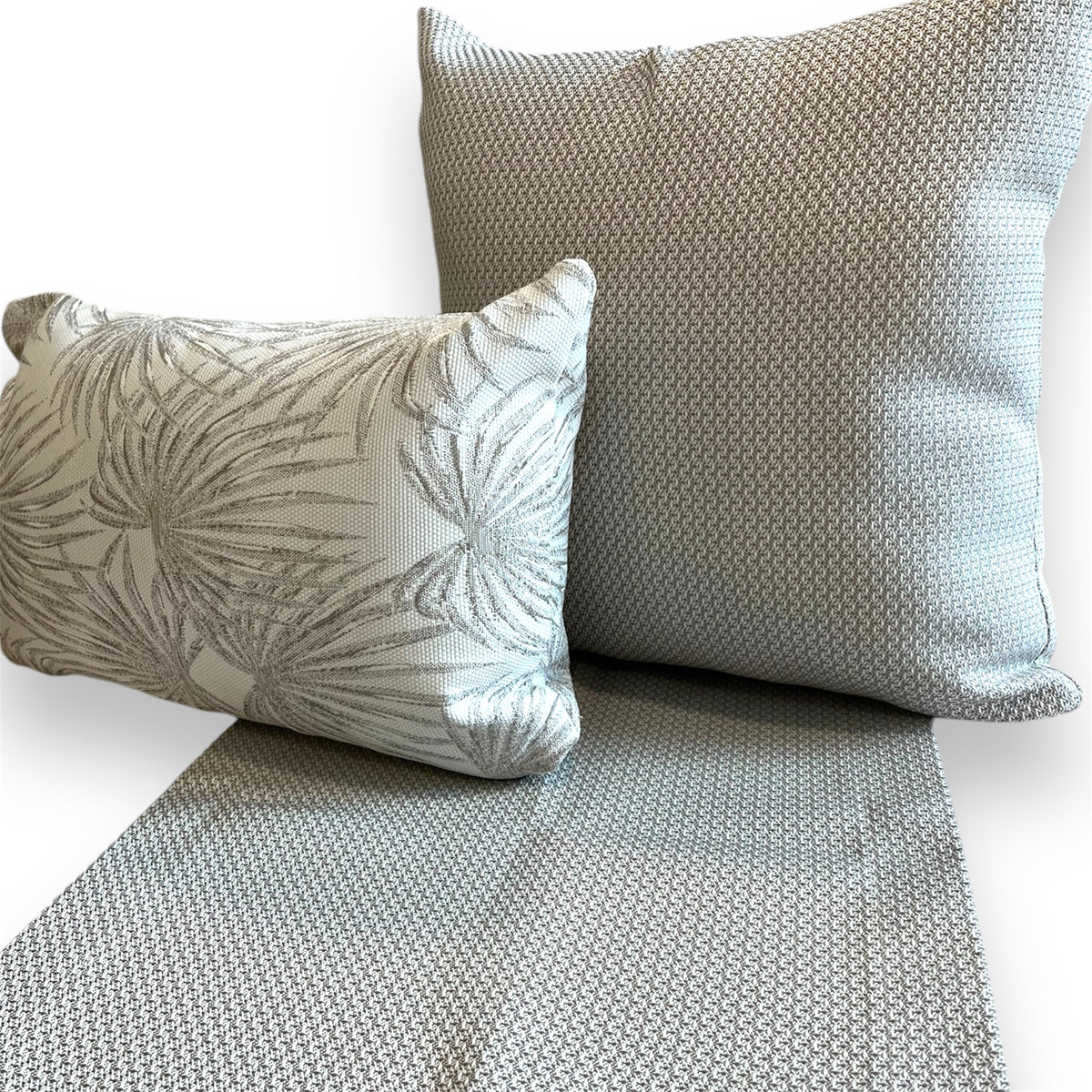 Textured grey woven table runner laid beneath a matching square throw pillow and a smaller rectangular tropical palm print pillow against a plain background.