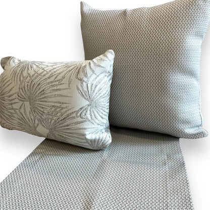 Textured grey woven table runner laid beneath a matching square throw pillow and a smaller rectangular tropical palm print pillow against a plain background.