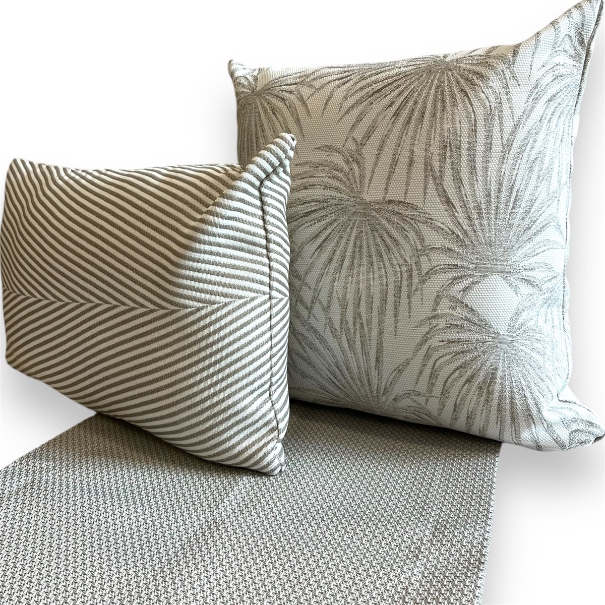 Pair of grey accent pillows—one smaller rectangular pillow with striped chevron stitching and one larger square pillow with a palm leaf pattern—resting on a matching textured woven grey runner.