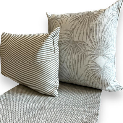 Pair of grey accent pillows—one smaller rectangular pillow with striped chevron stitching and one larger square pillow with a palm leaf pattern—resting on a matching textured woven grey runner.