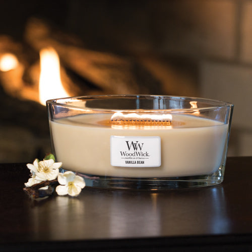 Lit WoodWick Vanilla Bean Ellipse Jar Candle glowing on a dark wooden surface with white orchid flowers and vanilla pods beside it, set against a warm fireplace background.