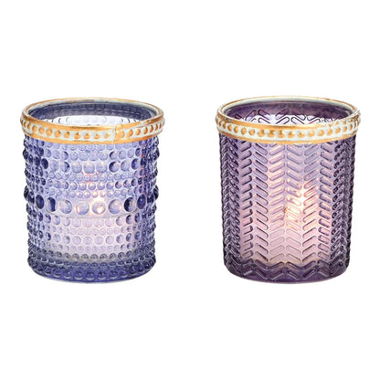 Two cylindrical glass candle holders with geometric patterns and brown rims, one with a purple hue and the other with a transparent appearance, both containing lit candles.