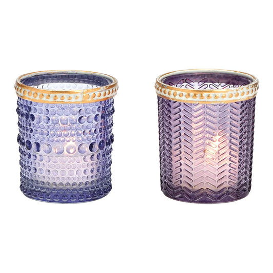 Two cylindrical glass candle holders with geometric patterns and brown rims, one with a purple hue and the other with a transparent appearance, both containing lit candles.