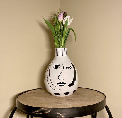 Decorative vase with face design on a wooden stool against a beige wall