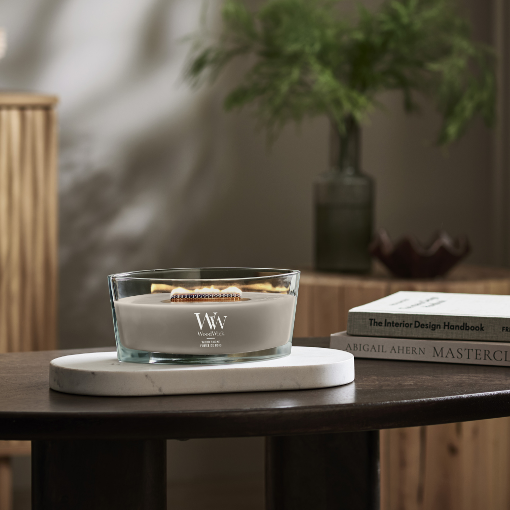 WoodWick Wood Smoke ellipse jar candle sitting on a marble trivet on a dark wooden table beside books.