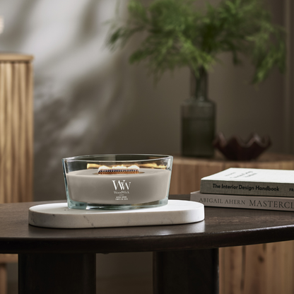 WoodWick Wood Smoke ellipse jar candle sitting on a marble trivet on a dark wooden table beside books.