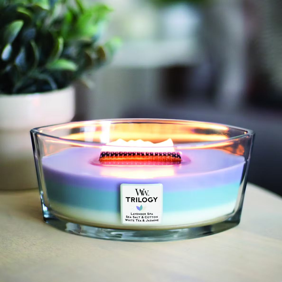 Lit WoodWick Calming Retreat Trilogy Ellipse Jar Candle with glowing flame, showcasing layered purple, blue, and white wax with the label listing Lavender Spa, Sea Salt & Cotton, and White Tea & Jasmine.