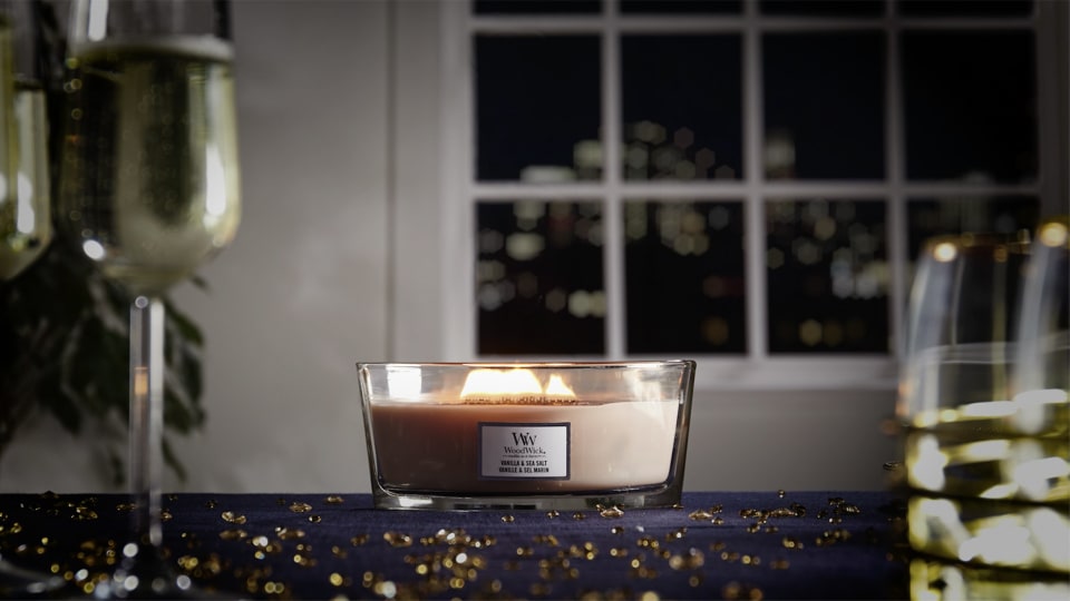 Lit WoodWick Ellipse Jar Candle glowing on a dark blue tablecloth scattered with gold glitter, flanked by champagne flutes in a festive holiday setup.